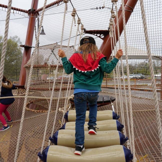 Children playing on a rope and log bridge at a playground, with one child balancing on cylindrical beams and another climbing a rope net.