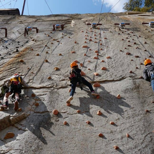 Three climbers ascending an outdoor artificial rock climbing wall equipped with secured ropes and various holds.