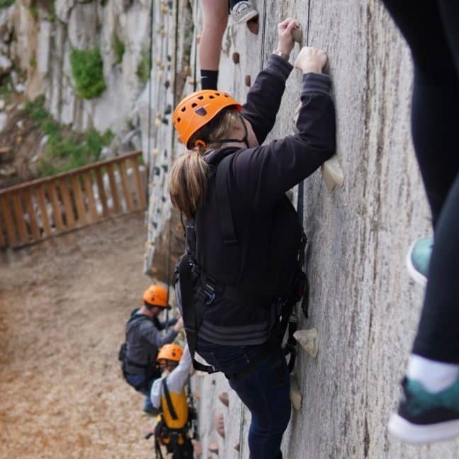 A climber wearing an orange helmet ascends a vertical, outdoor artificial climbing wall, gripping onto holds, with other climbers visible in the background.