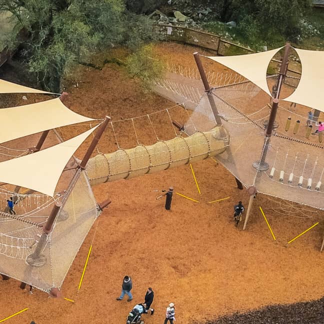 Aerial view of an outdoor playground with rope climbing structures and shaded areas under beige canopies, surrounded by trees. Several children and adults are present in groups.