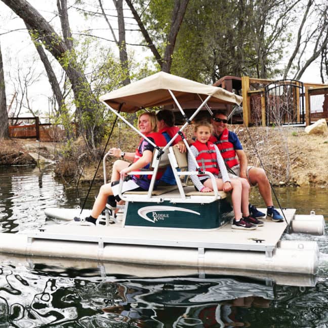 A family of four wearing life jackets riding a pedal boat on a calm lake, surrounded by trees.