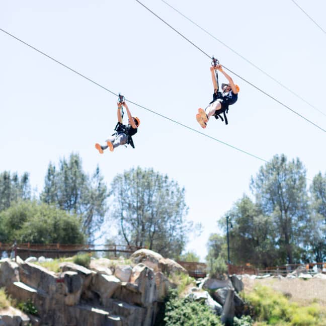 Two people ziplining over a rocky landscape with trees under a clear blue sky.